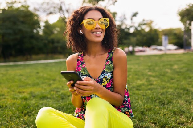 Молодая стильная улыбающаяся темнокожая женщина, использующая смартфон, слушая музыку на беспроводных наушниках, веселится в парке
