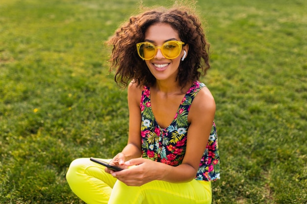 Young stylish smiling black woman using smartphone listening to music on wireless earphones having fun in park