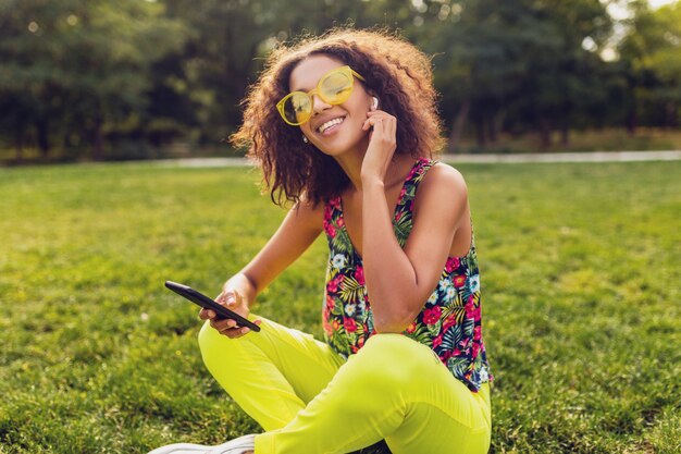 Молодая стильная улыбающаяся темнокожая женщина, использующая смартфон, слушая музыку на беспроводных наушниках, веселится в парке