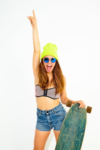 Giovane modello sorridente alla moda della donna in vestiti casuali dello swimwear di estate e berretto giallo che posano con lo scrittorio di longboard, mostrante il segno di rock-and-roll. isolato su bianco