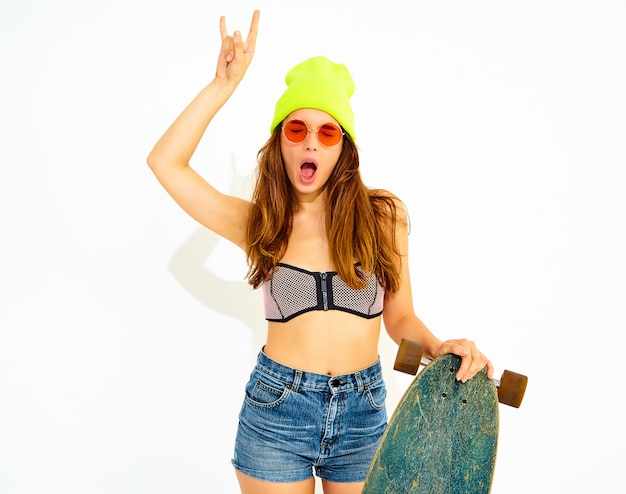 Young stylish smiling woman model in casual summer swimwear clothes and yellow beanie posing with longboard desk, showing rock and roll sign. Isolated on white