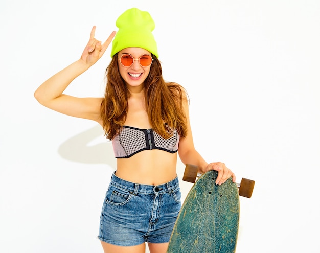 Young stylish smiling woman model in casual summer swimwear clothes and yellow beanie posing with longboard desk, showing rock and roll sign. isolated on white