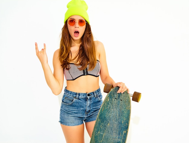 Young stylish smiling woman model in casual summer swimwear clothes and yellow beanie posing with longboard desk, showing rock and roll sign. Isolated on white