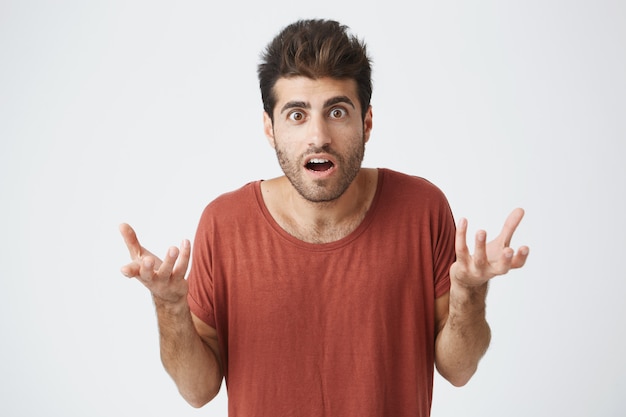 Young stylish spanish guy in red t-shirt with mouth wide opened, holding hands in surprised gesture shocked with his favourite football team loses in match. Body language