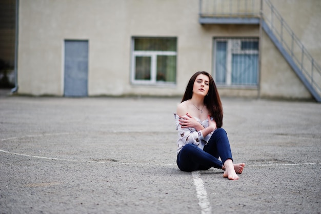 Young stylish teenage brunette girl on shirt pants and high heels shoes sitting on pavement and posed background school backyard Street fashion model concept