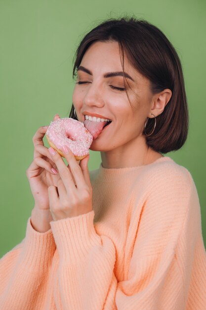 Young stylish woman in casual peach sweater  isolated on green olive wall licking pink donut copy space