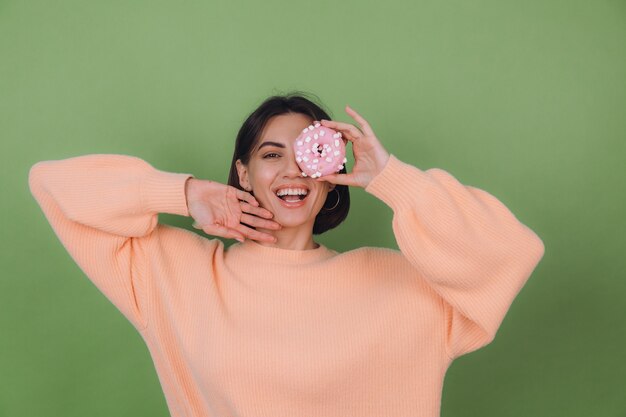Молодая стильная женщина в повседневном персиковом свитере изолирована на зеленой оливковой стене с розовым пончиком счастливая копия пространства