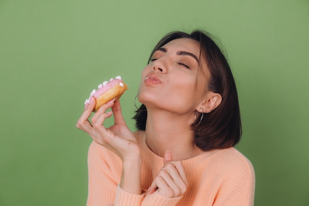 Young stylish woman in casual peach sweater and orange glasses isolated on green olive wall biting pink donut with closed eyes copy space