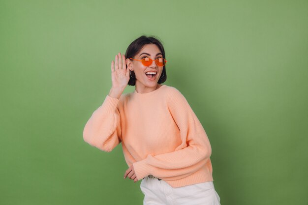 好奇心旺盛な緑のオリーブの壁に隔離されたカジュアルな桃のセーターとオレンジ色のメガネの若いスタイリッシュな女性は、耳のコピースペースで手であなたが言っていることを聞いてみてください