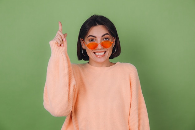 Young stylish woman in casual peach sweater and orange glasses isolated on green olive wall excited point index fingers up copy space