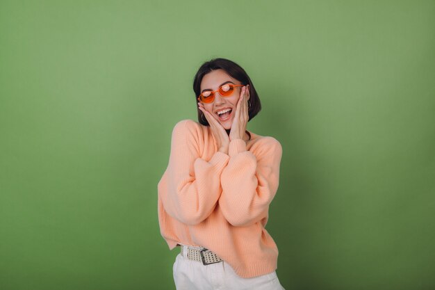 緑のオリーブの壁に分離されたカジュアルな桃のセーターとオレンジ色のメガネの若いスタイリッシュな女性