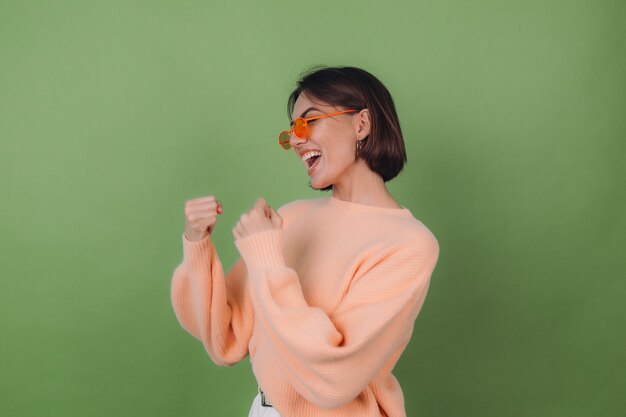 Young stylish woman in casual peach sweater and orange glasses isolated on green olive wall positive doing winner gesture clenching fist copy space