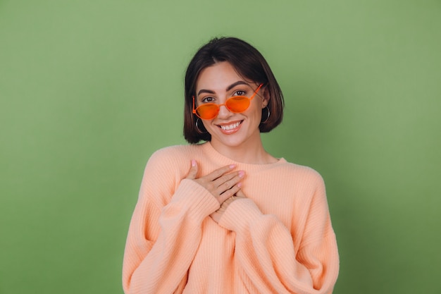 Молодая стильная женщина в повседневном персиковом свитере и оранжевых очках изолирована на зеленой оливковой стене, положительно держась за руки, сложенные на груди, пространство для копии сердца
