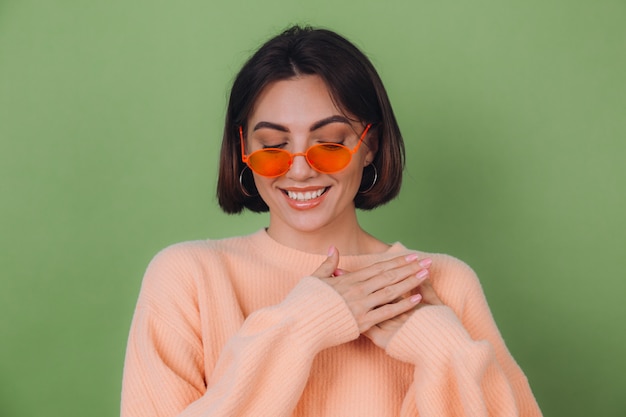 Young stylish woman in casual peach sweater and orange glasses isolated on green olive wall positive holding hands folded on chest, heart copy space