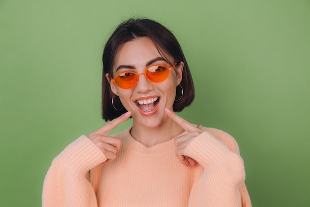 緑のオリーブの壁に分離されたカジュアルな桃のセーターとオレンジ色のメガネの若いスタイリッシュな女性人差し指で白い歯を指している肯定的な笑顔コピースペース