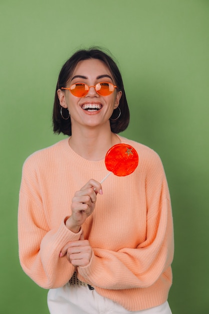 Young stylish woman in casual peach sweater and orange glasses isolated on green olive wall with orange lollipop positive smile copy space