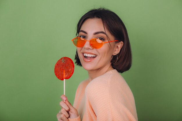 オレンジ色のロリポップのポジティブな笑顔のコピースペースと緑のオリーブの壁に分離されたカジュアルな桃のセーターとオレンジ色のメガネの若いスタイリッシュな女性