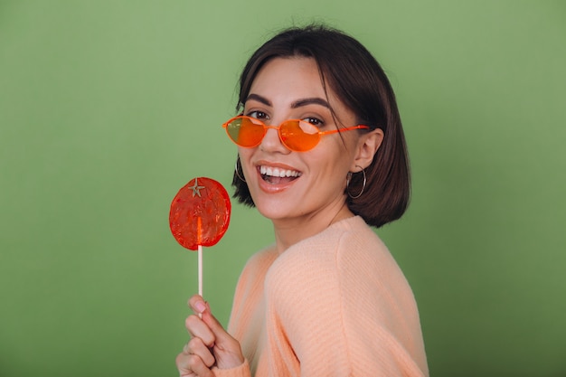 Young stylish woman in casual peach sweater and orange glasses isolated on green olive wall with orange lollipop positive smile copy space