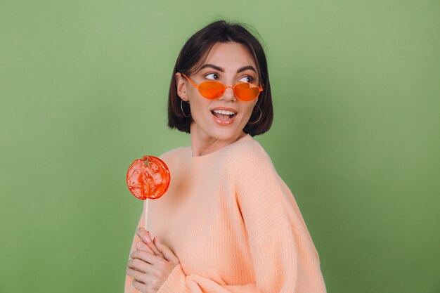 Молодая стильная женщина в повседневном персиковом свитере и оранжевых очках изолирована на зеленой оливковой стене с оранжевым леденцом на палочке позитивной улыбкой.