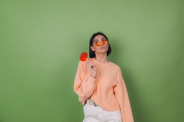 Молодая стильная женщина в повседневном персиковом свитере и оранжевых очках изолирована на зеленой оливковой стене с оранжевым леденцом вдумчивым взглядом в сторону, мысля копией пространства