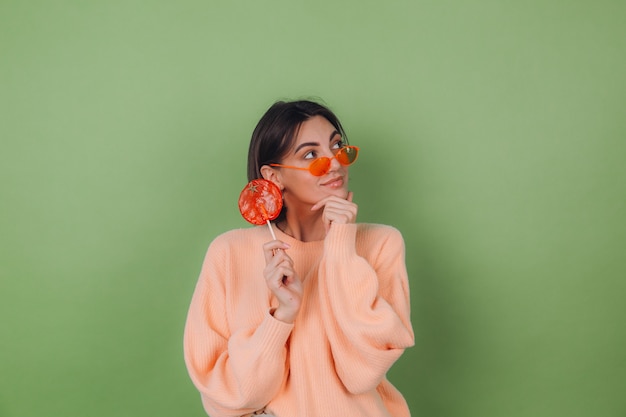 Young stylish woman in casual peach sweater and orange glasses isolated on green olive wall with orange lollipop thoughtful look aside thinking copy space