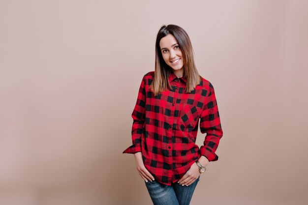 Young stylish woman dressed checkered shirt and jeans poses on the isolated wall with charming smile and happy emotions