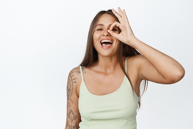 Young stylish woman laughing, showing okay sign against eye and smiling happy, standing in tank top against white background