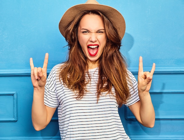 Young stylish woman model in casual summer clothes  and brown hat with red lips, posing near blue wall. Screaming and showing rock and roll sign