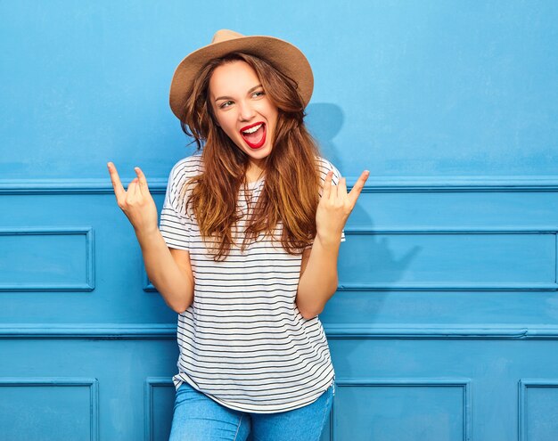 Молодая стильная модель женщины в повседневной летней одежде и коричневой шляпе с красными губами, позирующей около синей стены. Показывать знак рок-н-ролл