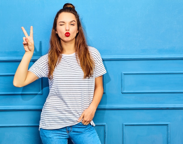 Young stylish woman model in casual summer clothes with red lips, posing near blue wall. Giving kiss and peace sign