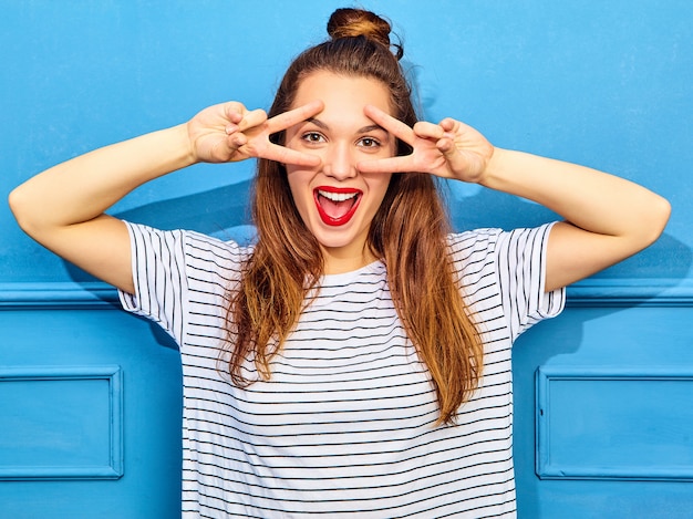 Free photo young stylish woman model in casual summer clothes with red lips, posing near blue wall. screaming and showing peace sign