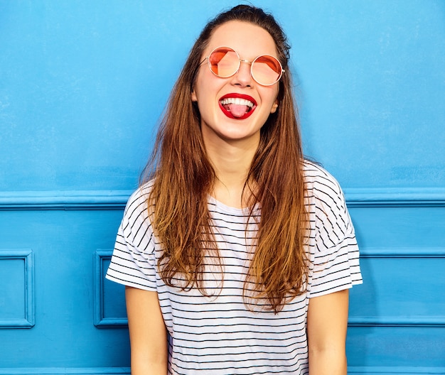 Модель молодой стильной женщины в повседневной летней одежде с красными губами, позирует возле синей стены. Показывая ее язык