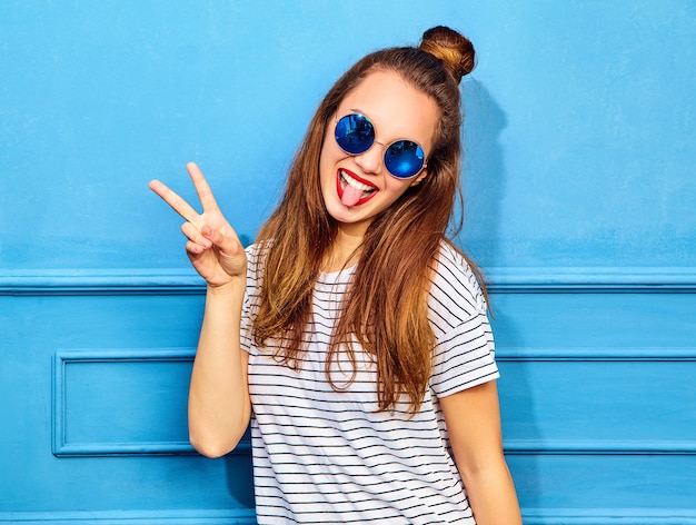 Free photo young stylish woman model in casual summer clothes with red lips, posing near blue wall. showing peace sign and her tongue