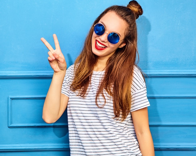 Young stylish woman model in casual summer clothes with red lips, posing near blue wall. Showing peace sign