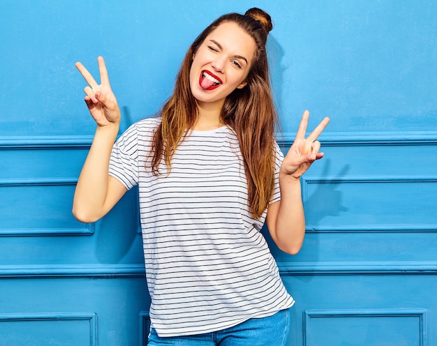Young stylish woman model in casual summer clothes with red lips, posing near blue wall. Winking and showing peace sign