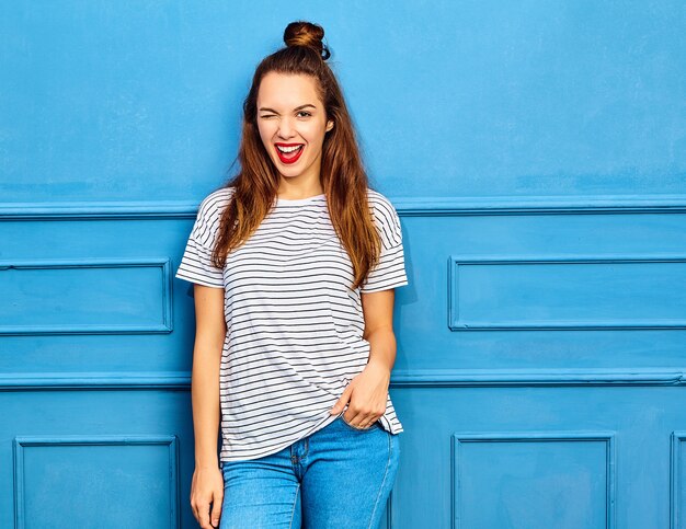 Young stylish woman model in casual summer clothes with red lips, posing near blue wall. Winking