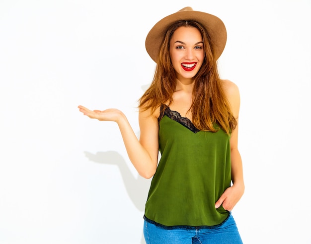 Молодая стильная модель женщины в повседневной летней зеленой одежде и коричневой шляпе с красными губами, держащей что-то на руке, изолировано