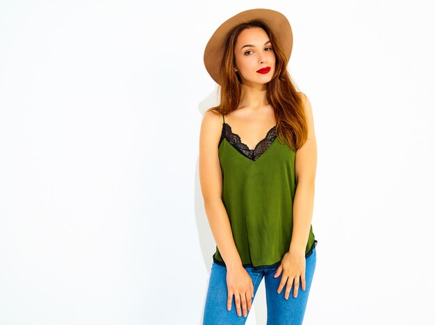 Молодая стильная модель женщины в непринужденной летней зеленой одежде и коричневой шляпе с красными губами, позирующей возле белой стены