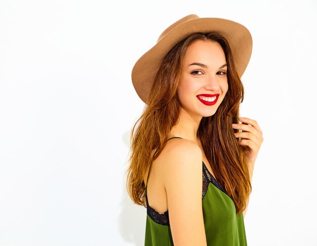 Young stylish woman model in casual summer green clothes and brown hat with red lips, posing near white wall