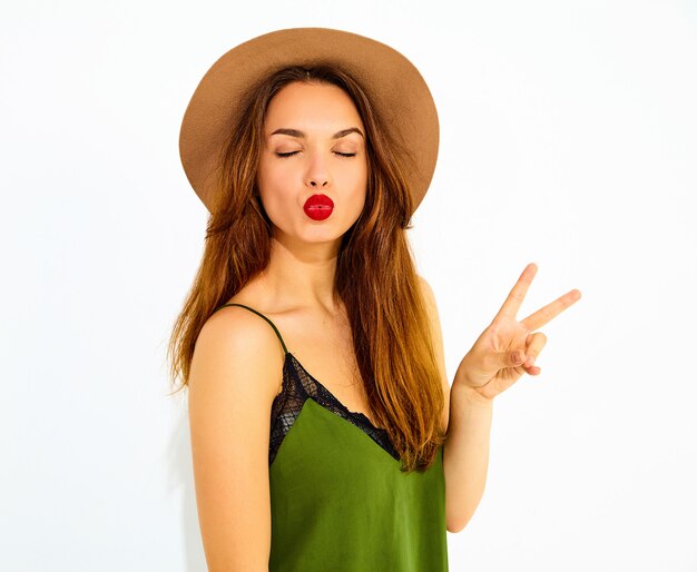 Молодая стильная модель женщины в повседневной летней зеленой одежде и коричневой шляпе с красными губами. Показывать знак мира и давать поцелуй