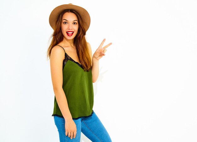 Young stylish woman model in casual summer green clothes and brown hat with red lips, showing peace sign and her tongue