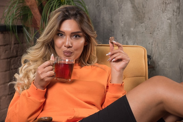 Young stylish woman sitting in armchair blowing cup of tea. 