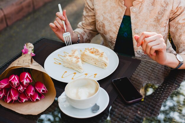 Young stylish woman sitting in cafe, holding cup of cappuccino, eating tasty cake