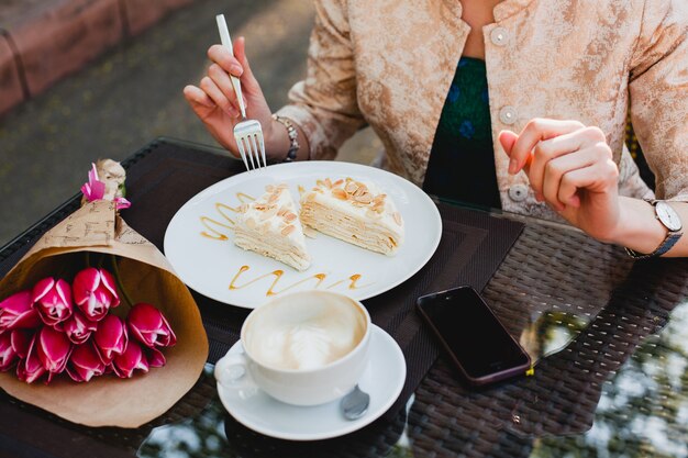 Молодая стильная женщина сидит в кафе, держит чашку капучино и ест вкусный торт