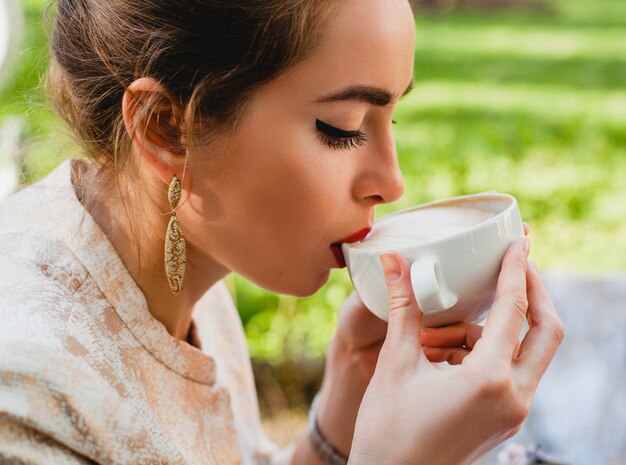 カプチーノのカップを保持しているカフェに座っている若いスタイリッシュな女性