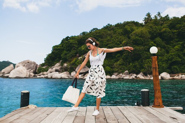 Young stylish woman standing on pier, walking, listening to music on headphones, summer apparel, white skirt, handbag, azure water, landscape background, tropical lagoon, vacation, traveling in asia