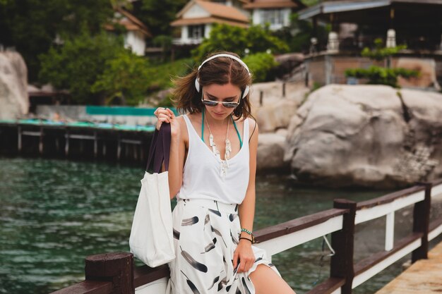 Young stylish woman standing on pier, walking, listening to music on headphones, summer apparel, white skirt, handbag, azure water, landscape background, tropical lagoon, vacation, traveling in asia