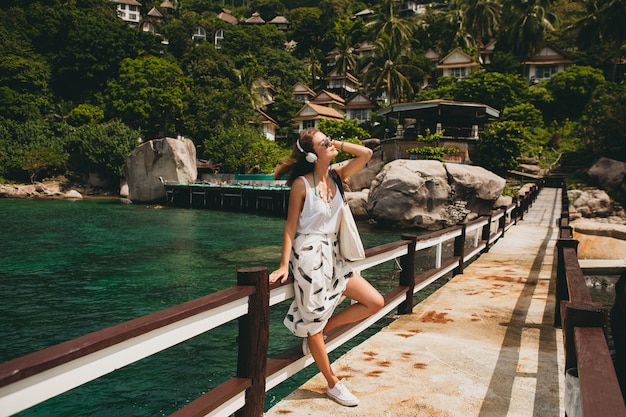 Young stylish woman standing on pier, walking, listening to music on headphones, summer apparel, white skirt, handbag, azure water, landscape background, tropical lagoon, vacation, traveling in asia