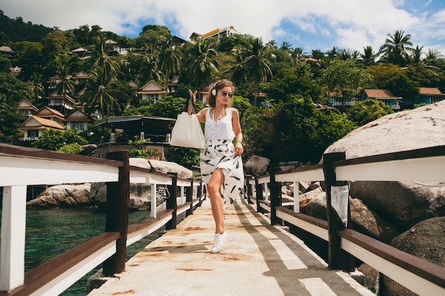 Young stylish woman standing on pier, walking, listening to music on headphones, summer apparel, white skirt, handbag, azure water, landscape background, tropical lagoon, vacation, traveling in asia