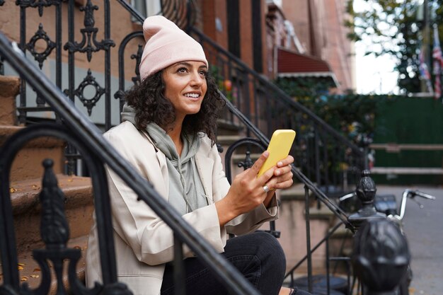 Young stylish woman using smartphone while sitting on steppes in the city